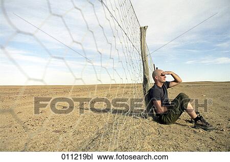 Stock Image - man sitting by a fence in the desert. Fotosearch