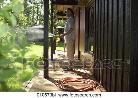 Stock Image - man standing on veranda of weekend home watering the lawn. Fotosearch