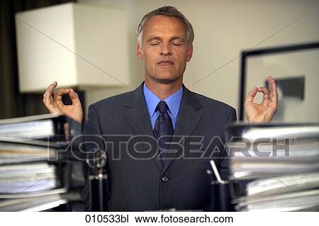 Stock Image - mature businessman in meditation pose sitting behind piles of folders. Fotosearch