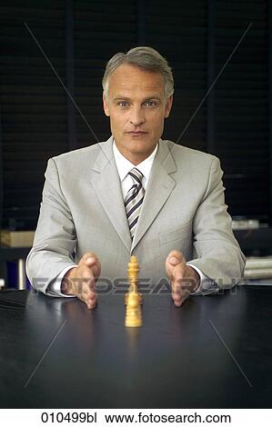 Stock Image - mature businessman sitting at desk and making explanations with chess pieces. Fotosearch
