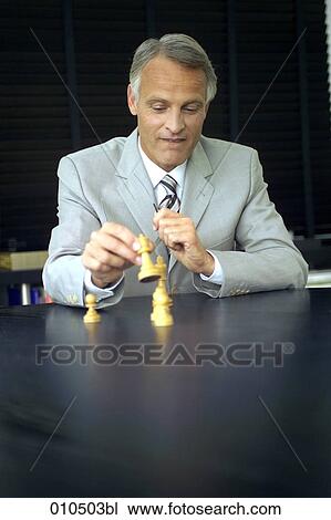 Stock Image - mature businessman sitting at desk and playing with chess pieces. Fotosearch