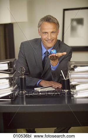 mature businessman sitting at his desk and holding a little globe in his hand View Large Photo Image Stock Image - mature businessman sitting at his desk and holding a little globe in his hand. Fotosearch