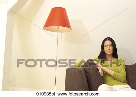 Stock Image - Portrait of young woman in casual dress sitting on a sofa. Fotosearch