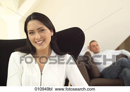 Smiling young woman sitting in a chair with boyfriend in the background View Large Photo Image Stock Image - Smiling young woman sitting in a chair with boyfriend in the background. Fotosearch
