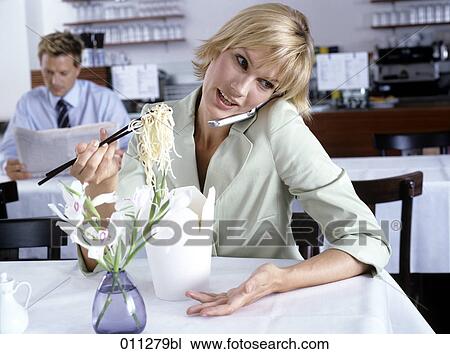 Stock Image - woman eating noodles while talking on mobile; businessman reading paper in the background. Fotosearch