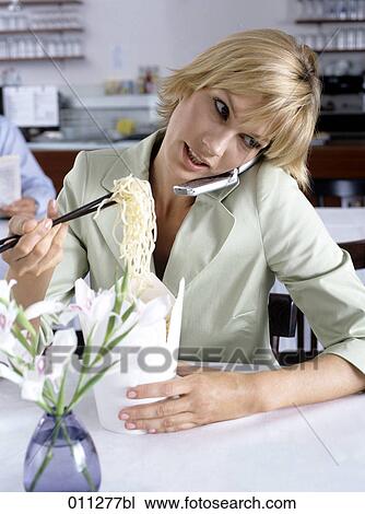 Stock Image - woman eating noodles with chopsticks while talking on mobile. Fotosearch