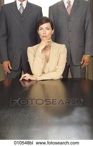 young businesswoman sitting at table with two businessmen standing in the background View Large Photo Image Stock Image - young businesswoman sitting at table with two businessmen standing in the background. Fotosearch