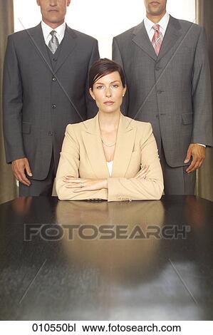 young businesswoman sitting at table with two businessmen standing in the background View Large Photo Image Stock Image - young businesswoman sitting at table with two businessmen standing in the background. Fotosearch