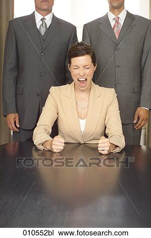 young businesswoman sitting at table and shouting while two businessmen are standing in the background View Large Photo Image Stock Image - young businesswoman sitting at table and shouting while two businessmen are standing in the background. Fotosearch