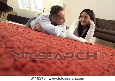 Stock Image - Young couple lying on the carpet of their living room looking at each other. Fotosearch