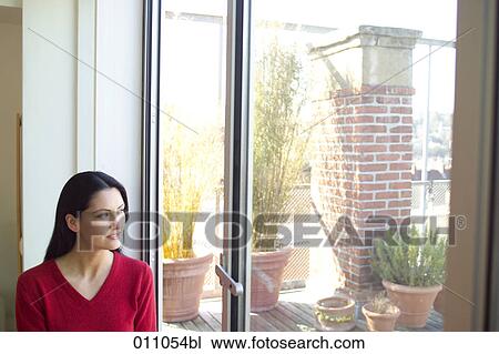 Stock Image - Young woman looking through window. Fotosearch