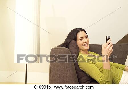 Stock Image - Young woman lying on sofa looking at display of her mobile phone. Fotosearch