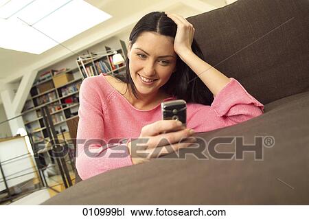 Young woman resting on sofa looking at display of mobile phone View Large Photo Image Stock Image - Young woman resting on sofa looking at display of mobile phone. Fotosearch