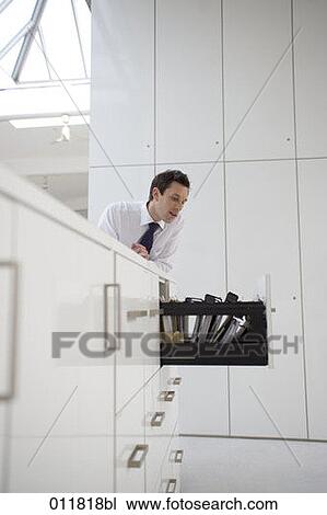 Stock Image - businessman leaning on filing cabinet looking at open drawer. Fotosearch