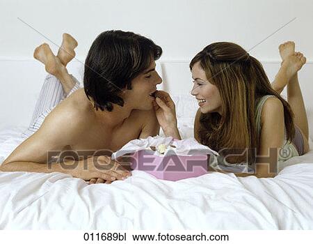 Stock Image - couple lying on bed, eating chocolates. Fotosearch