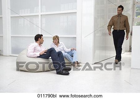 Stock Image - man passing by couple on bean bags in office building. Fotosearch