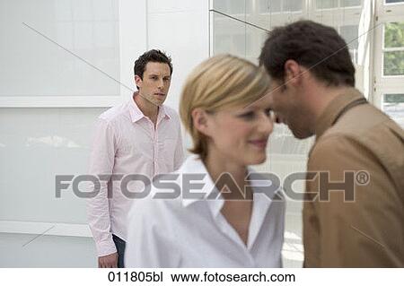 Stock Image - office workers whispering to each other, uneasy looking man listening in the background. Fotosearch