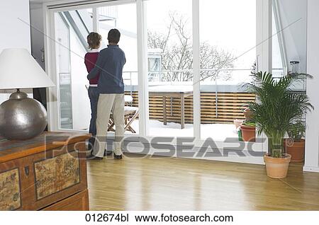 Stock Image - mature couple looking out of the window of their living room. Fotosearch