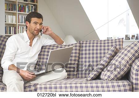 Stock Image - mature male sitting couch with mobile phone and laptop. Fotosearch