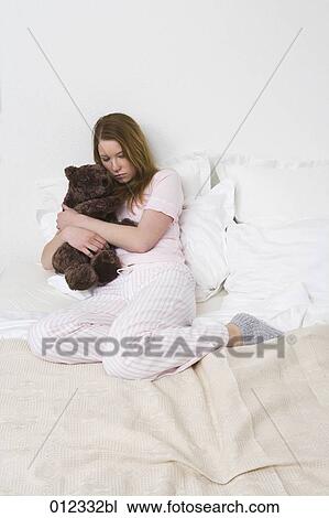 Stock Image - sad teenager sitting on bed and hugging teddy bear. Fotosearch