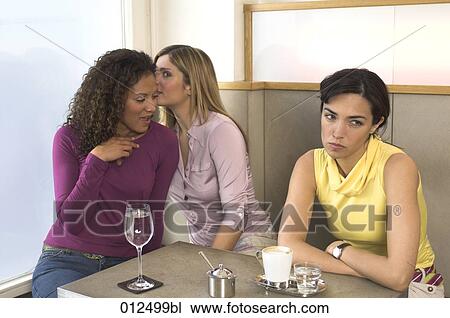 Stock Image - two female friends whispering to each other while third woman is looking enervated. Fotosearch