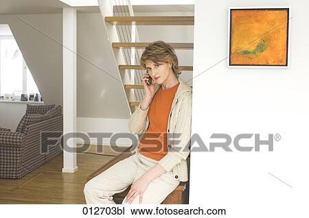 Stock Image - worried looking woman sitting under stairs at home and talking on her mobile. Fotosearch