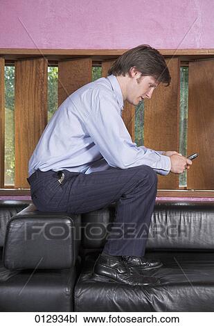Stock Image - businessman sitting on top of sofa checking his cellphone for text messages. Fotosearch