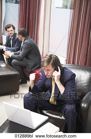 Stock Image - determined looking businessman sitting in front of notebook talking on his mobile. Fotosearch