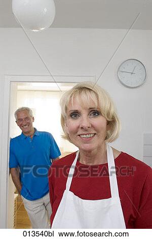 Stock Image - mature woman in kitchen with husband laughing in the background. Fotosearch