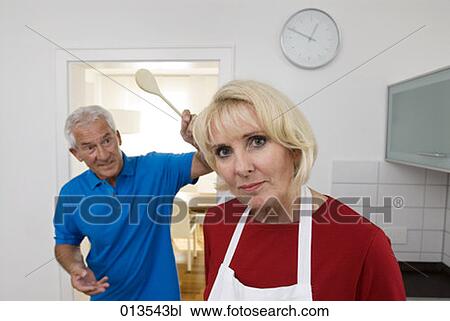 Stock Image - portrait of woman in kitchen with man trying to beat her playfully with wooden spoon. Fotosearch