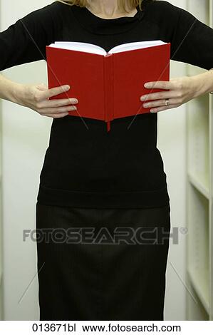 Stock Image - woman standing between shelves reading book. Fotosearch