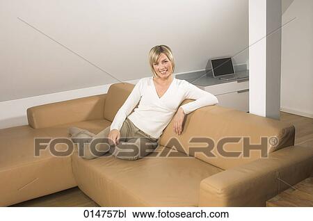 Stock Image - full body portrait of smiling young woman sitting on leather couch in living room. Fotosearch