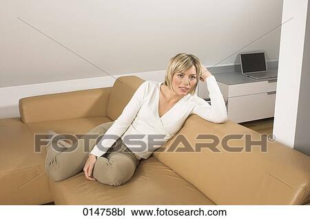 Stock Image - full body portrait of young woman sitting on leather couch in living room. Fotosearch