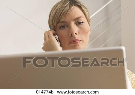 Stock Image - headshot of young woman holding mobile phone and looking at screen of her laptop computer. Fotosearch