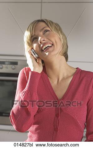 Stock Image - portrait of laughing young woman with cellphone in kitchen. Fotosearch