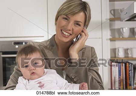Stock Image - portrait of smiling young businesswoman in kitchen holding baby and talking on mobile phone. Fotosearch