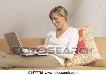 Stock Image - portrait of smiling young woman sitting on couch and working with notebook computer. Fotosearch