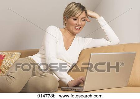 Stock Image - portrait of smiling young woman sitting on couch and working with notebook computer. Fotosearch