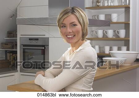 Stock Image - portrait of smiling young woman standing in kitchen. Fotosearch