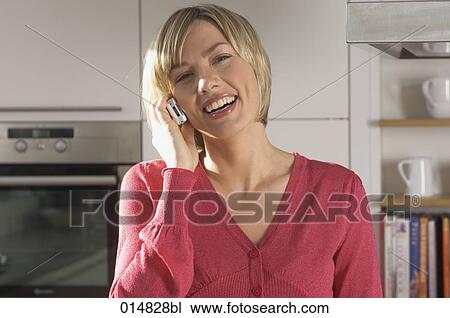 Stock Image - portrait of smiling young woman with cellphone in kitchen. Fotosearch