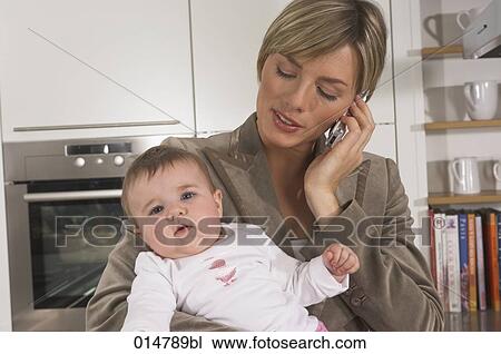 Stock Image - portrait of young businesswoman in kitchen holding baby and talking on mobile phone. Fotosearch
