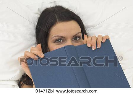 Stock Image - portrait of young woman in bed hiding her face behind book. Fotosearch