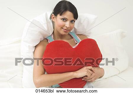portrait of young woman lying in bed holding red cushion in shape of heart View Large Photo Image Stock Image - portrait of young woman lying in bed holding red cushion in shape of heart. Fotosearch