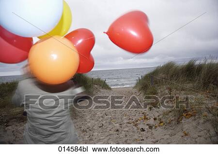 Stock Image - rear view of woman on windy beach in autumn holding colourful balloons. Fotosearch