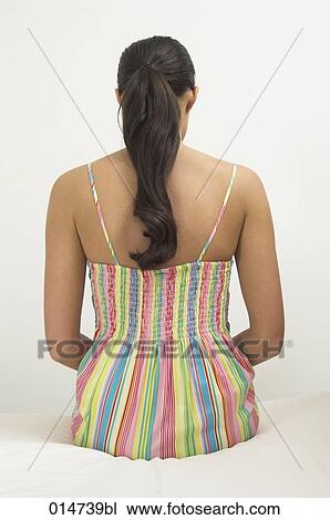 Stock Image - rear view of young woman with ponytail sitting on bed in colourful summer dress. Fotosearch