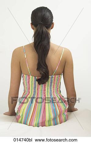 Stock Image - rear view of young woman with ponytail sitting on bed in colourful summer dress. Fotosearch