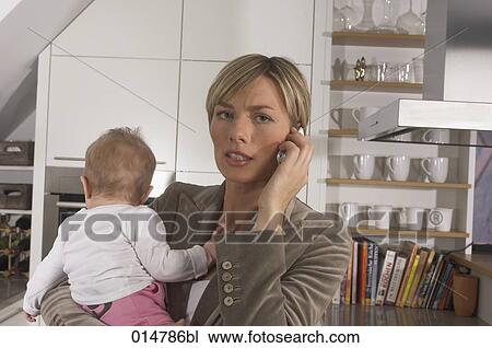 Stock Image - stressed young businesswoman standing in kitchen holding baby and talking on mobile phone. Fotosearch