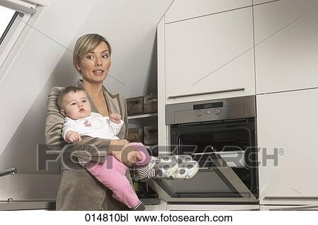 Stock Image - young businesswoman holding her baby while standing at the oven. Fotosearch
