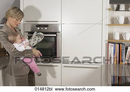Stock Image - young businesswoman holding her baby while standing at the oven. Fotosearch