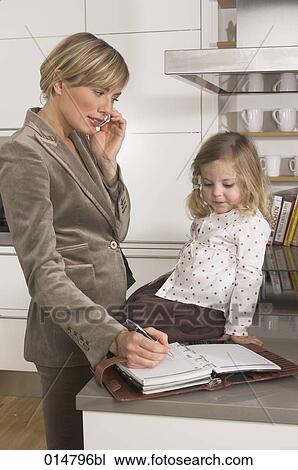 Stock Image - young businesswoman in kitchen looking at her daughter while listening to cellphone. Fotosearch